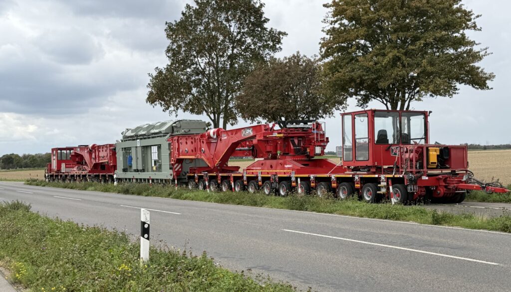 Transformator in der roten Scherenhubbrücke G|K400 der KAHL Unternehmensgruppe auf mehrachsigen Schwerlastmodulen, seitlich an einer Landstraße aufgenommen.