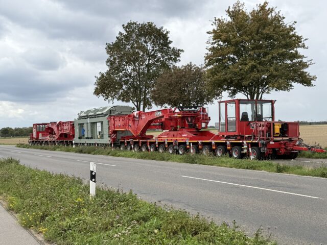 Transformator in der roten Scherenhubbrücke G|K400 der KAHL Unternehmensgruppe auf mehrachsigen Schwerlastmodulen, seitlich an einer Landstraße aufgenommen.