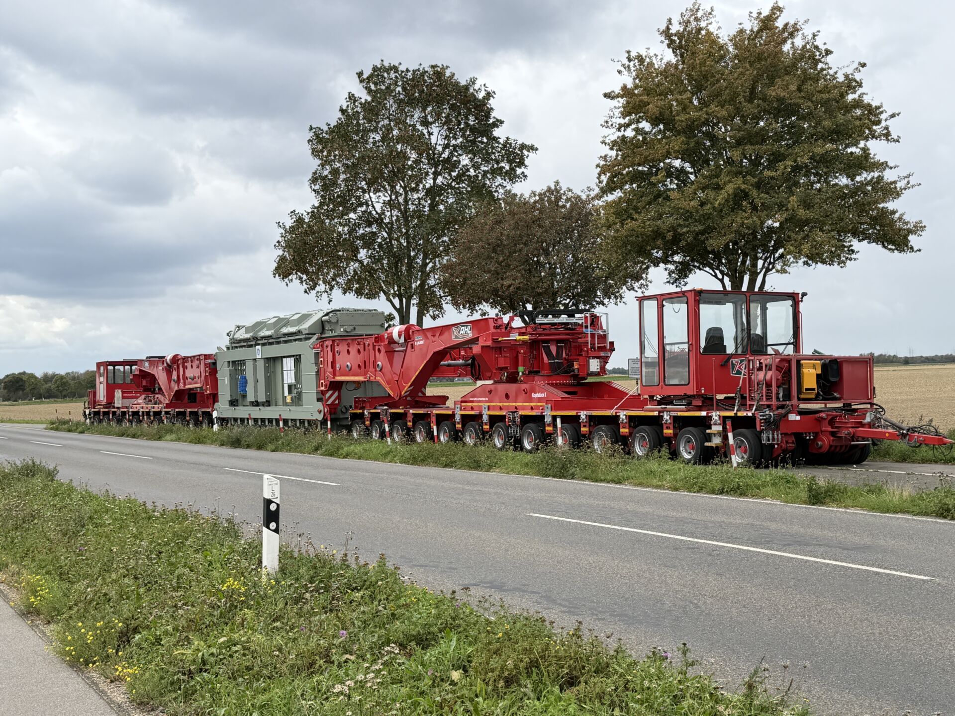 Transformator in der roten Scherenhubbrücke G|K400 der KAHL Unternehmensgruppe auf mehrachsigen Schwerlastmodulen, seitlich an einer Landstraße aufgenommen.